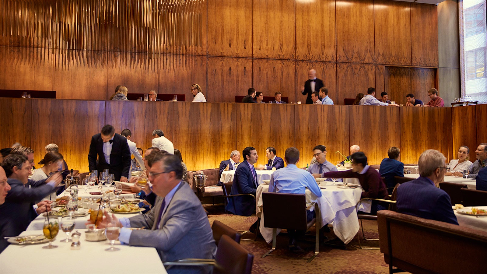 Diners in the Grill’s contemporary-design dining room with plush, fabric booths and metal-leather chairs, geometric, gold-colored chandeliers above and a large window looking onto a neighboring building